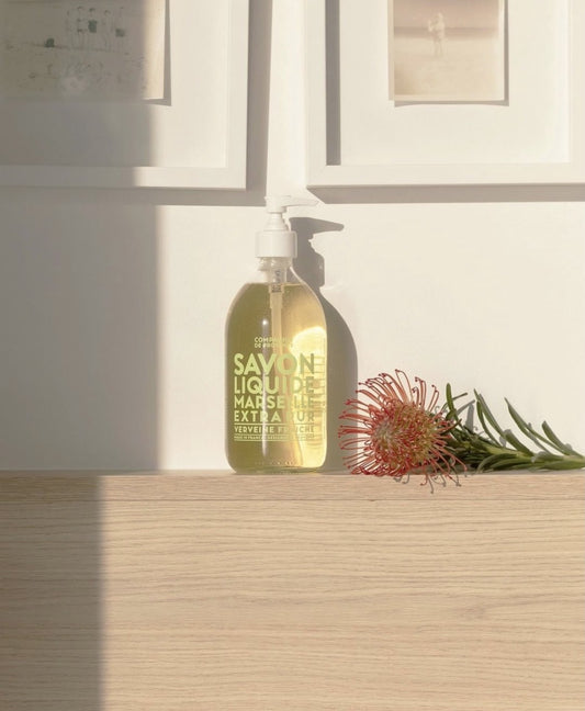 Compagnie de Provence Fresh Verbena liquid soap on an oak plinth with a protea, window shadow on the wall